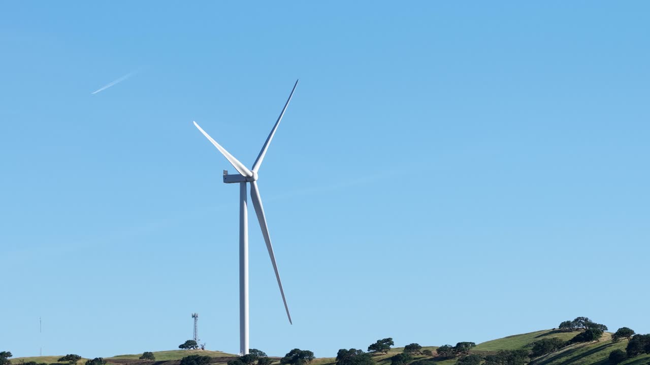 White wind turbine generating clean, renewable energy with its blades spinning against a bright blue sky, set on a green hill with a telecommunication tower in the background