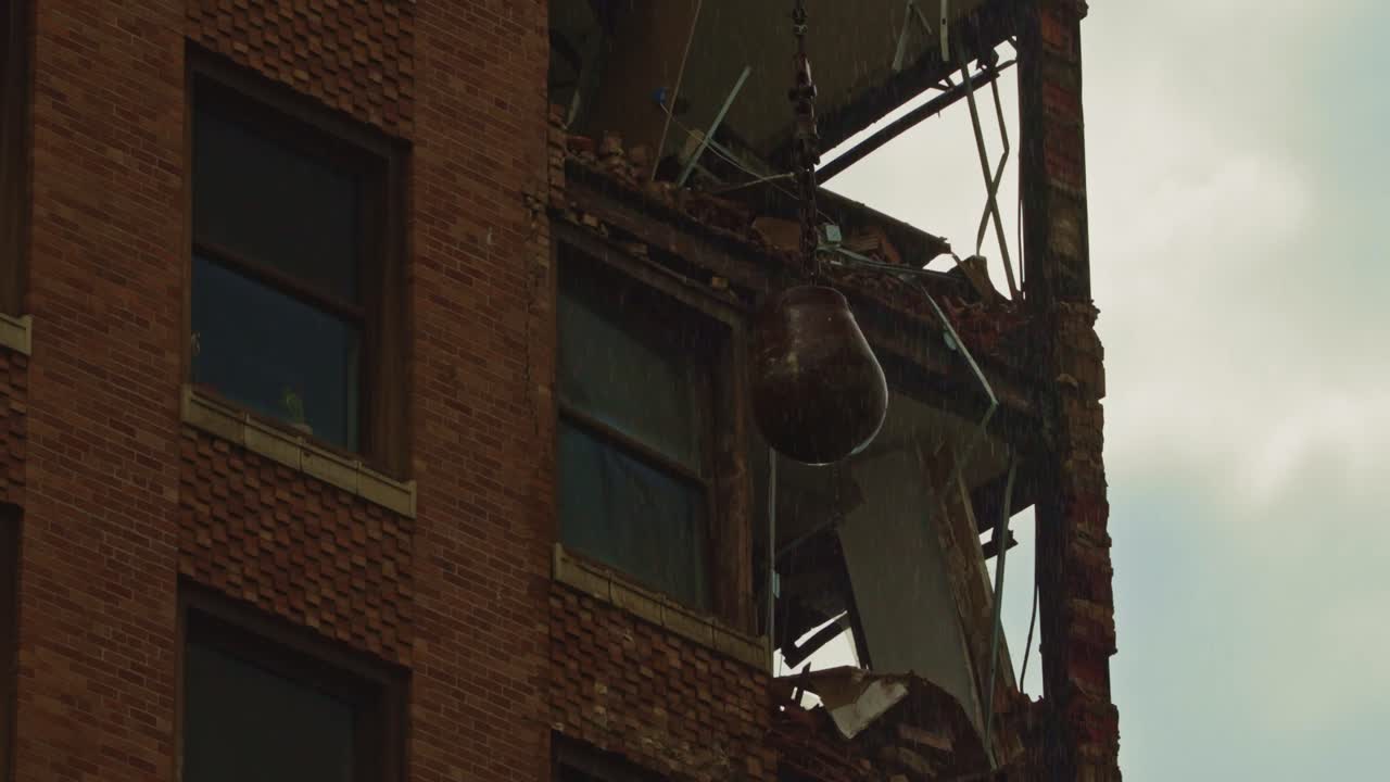 A heavy wrecking ball slams into the upper floor of a downtown brick building during controlled demolition, revealing crumbling structure and debris.