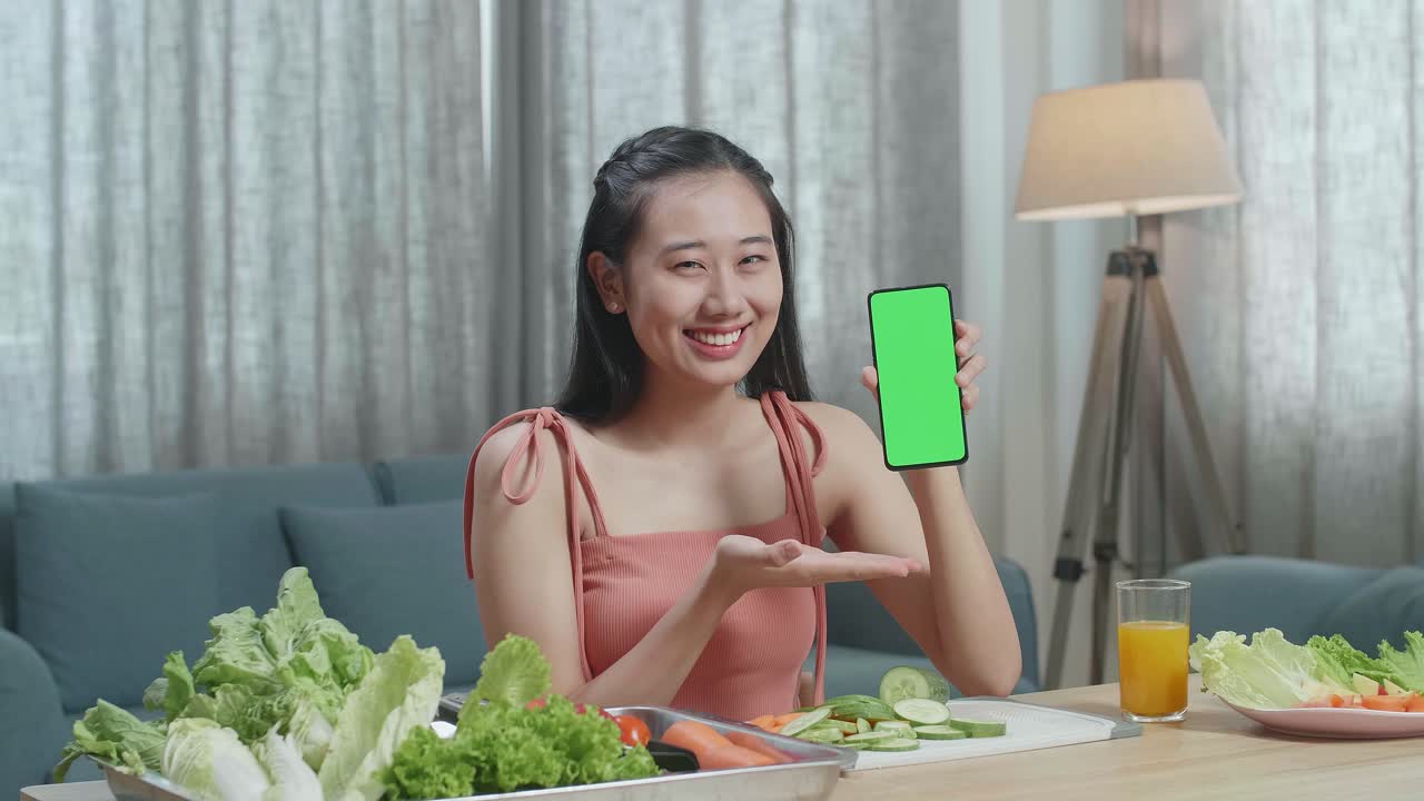 mujer asiática con comida saludable sosteniendo y señalando un teléfono inteligente de pantalla verde mientras está sentada en casa