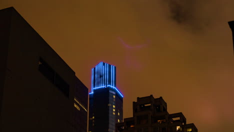 Stadtnacht-Mit-Orangefarbenen-Wolken,-Lichtverschmutzung,-Die-An-Einem-Hohen-Gebäude-Vorbeizieht,-Das-Mit-Blauen-LEDs-Im-Zeitraffer-Beleuchtet-Ist