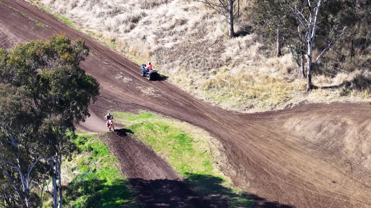 Motocross rider speeds around dirt track curve, aerial view, bright daylight, natural outdoor setting