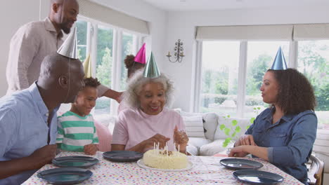 multi generation family sit around table celebrating grandmother's birthday as she blows out candles