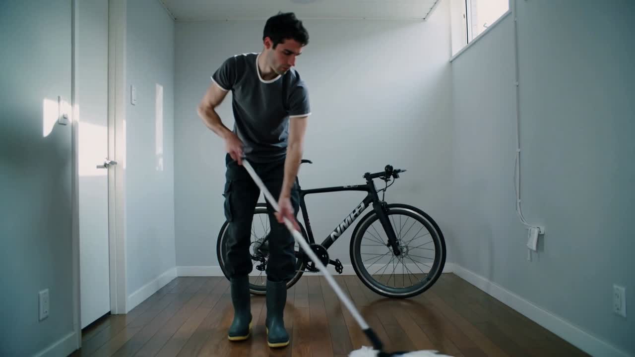 Starting with forward mop stroke, man in grey T-shirt sweeping room at home around road bicycle
