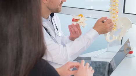 using a model of a spine, the doctor is sharing information with the patient, gesturing with their thumb and elbow to explain a medical condition