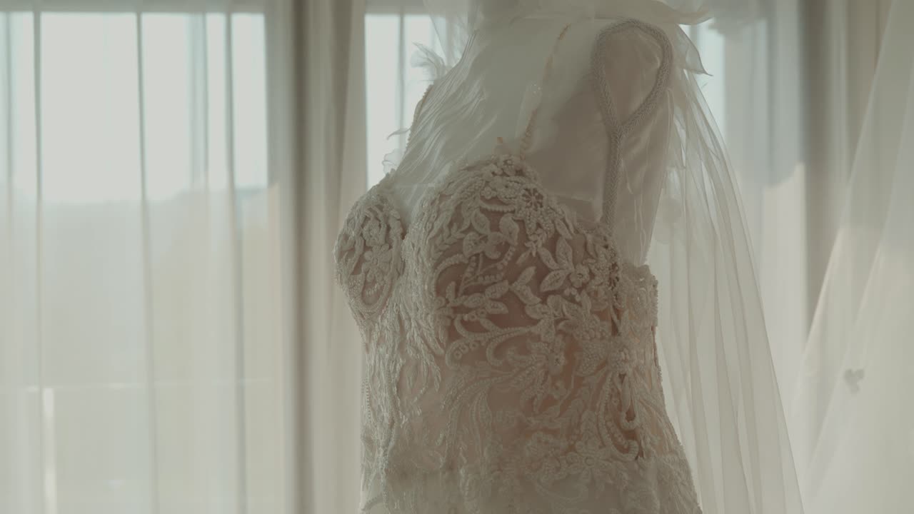 Изящное кружевное свадебное платье висит рядом с прозрачными шторами в теплом свете