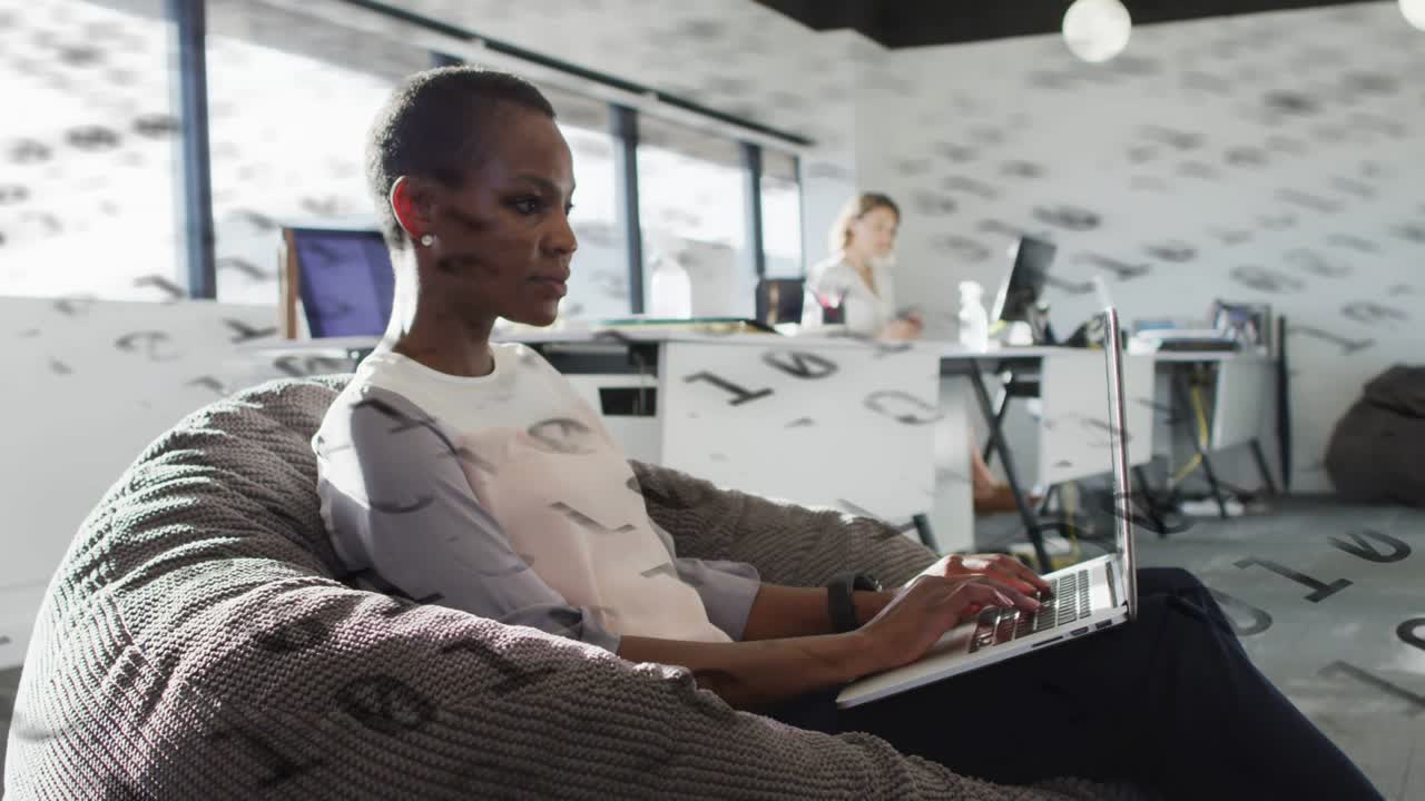 Mid-adult woman typing on laptop in beanbag, activating drifting binary overlay for technology demo
