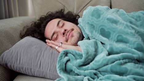 turn on the guy with curly hair and a mustache lies on a gray pillow, wrapped in a blue blanket and sleeps on the sofa at home