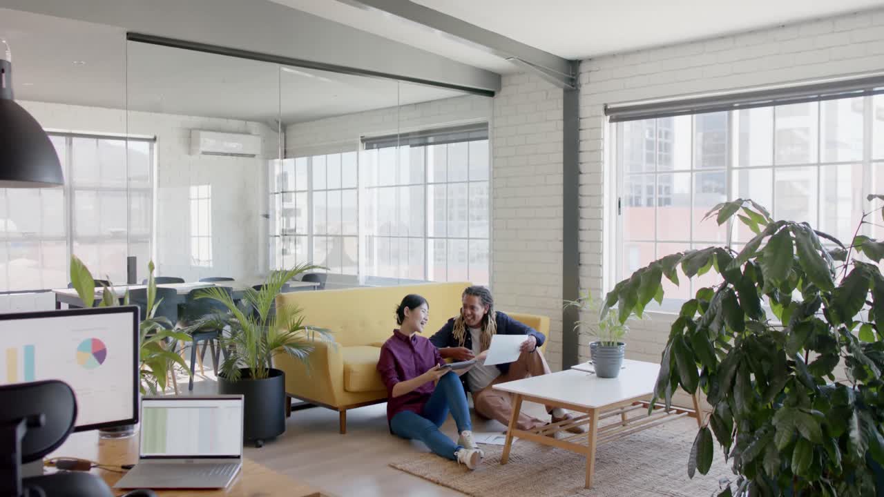 felices colegas creativos diversos usando tableta y discutiendo sentados en el piso de la oficina, cámara lenta