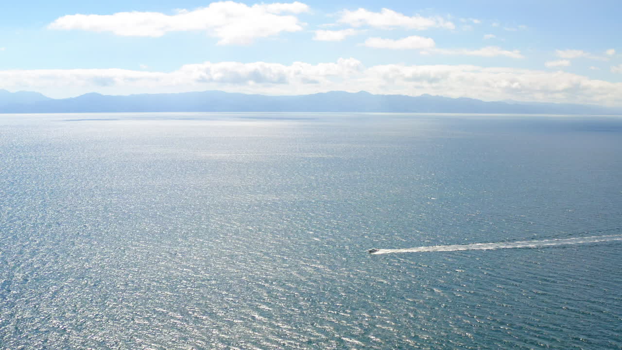 A boat creating a wake on a vast body of water with mountains and clouds in the distance