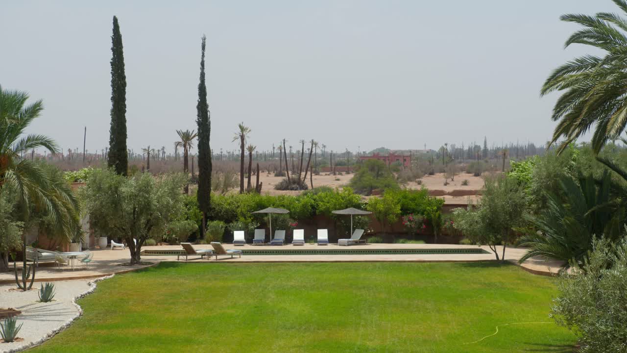 Wide static shot showcasing a vibrant green garden and swimming pool surrounded by desert terrain in Marrakesh, Morocco, highlighting contrast between nature and arid environment