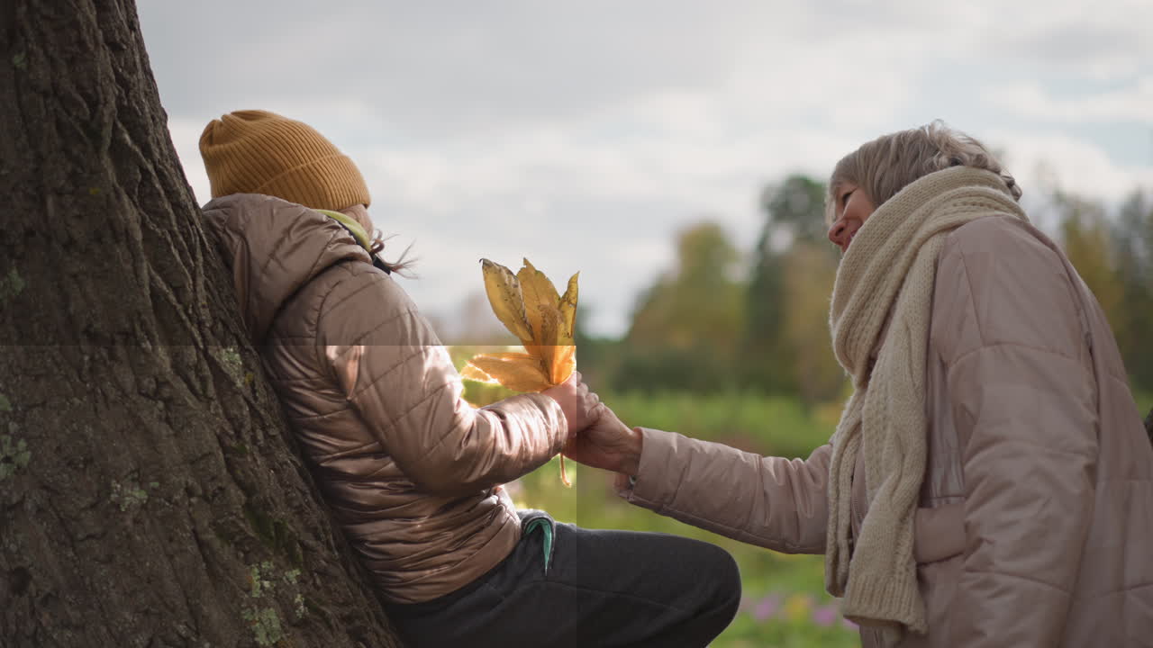 mom walks to daughter resting on tree trunk and hands over autumn leaf with gentle peck on cheek in peaceful fall park filled with colorful foliage and soft cloudy sky in background