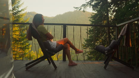 mujer disfruta de la atmósfera de la naturaleza matutina sola. mujer relajada se sienta en una silla en la terraza rodeada de un ambiente tranquilo de verano de observación del bosque