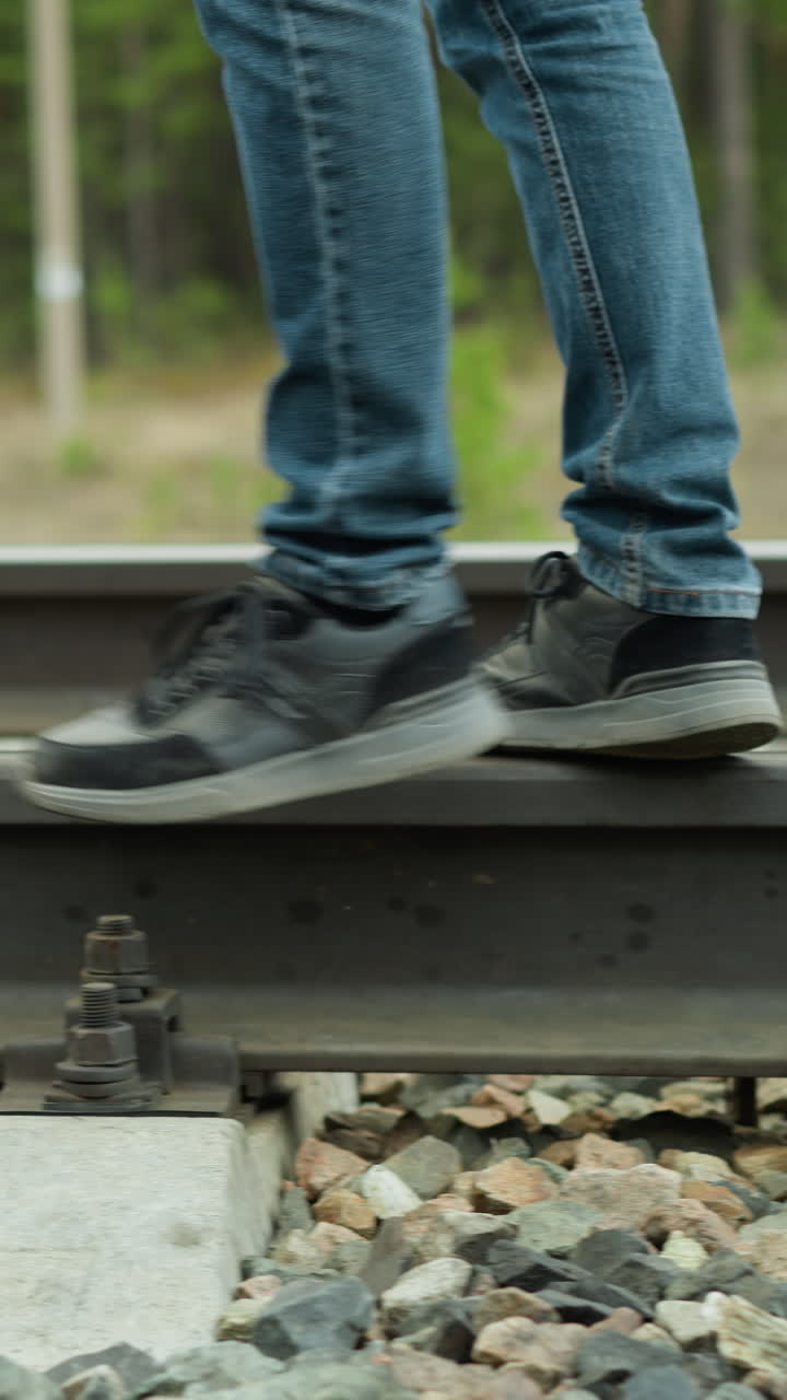 una vista de cerca de las piernas de un hombre en vaqueros azules y zapatillas negras caminando a lo largo de las vías del ferrocarril, el tiro se centra en los pies