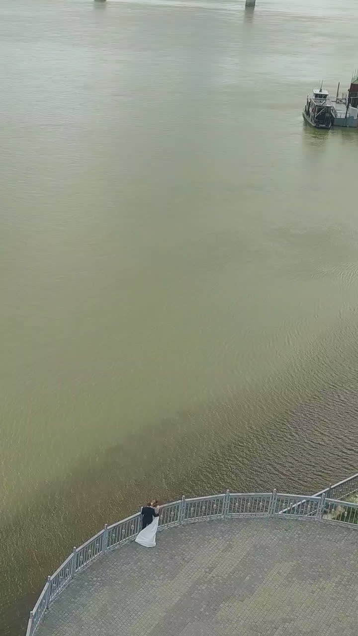 just married bride and groom stand on empty viewpoint at waterfront over wide calm river on autumn day aerial view