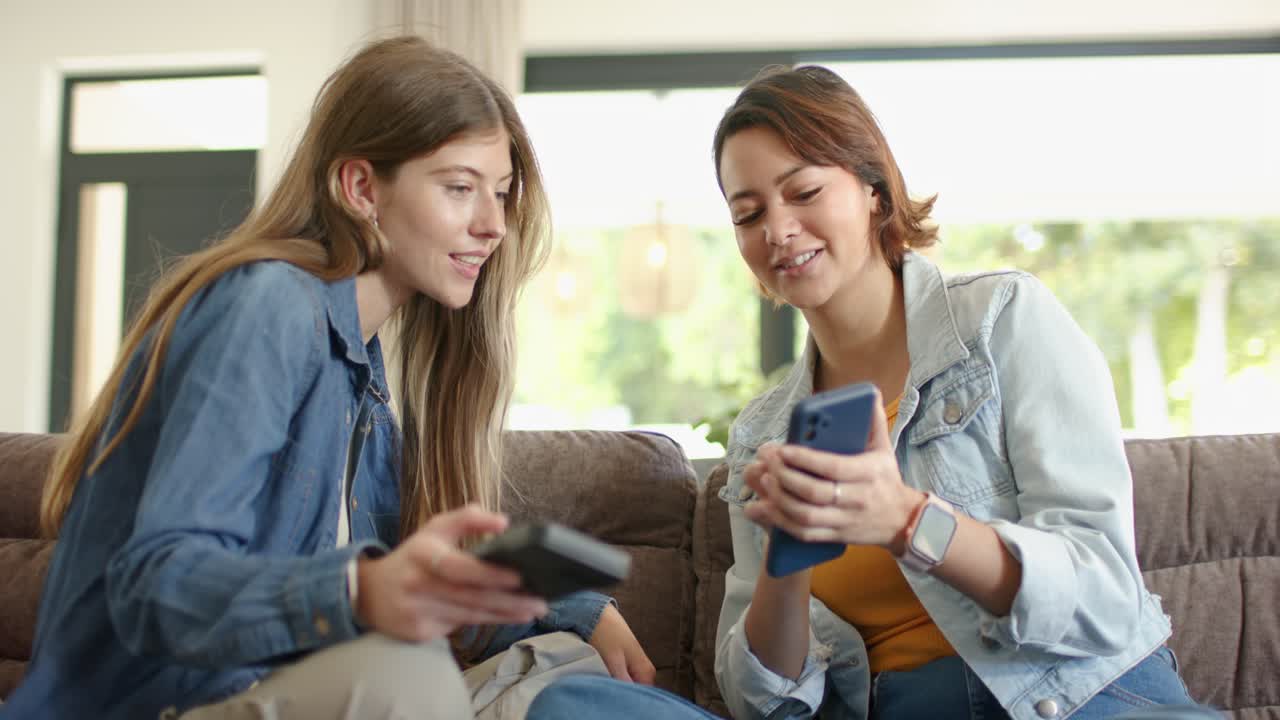 Female friends sharing phone on sofa by window, right woman tapping while left woman placing remote
