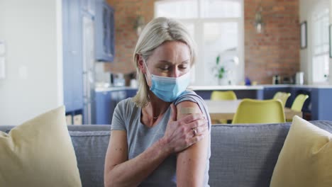 Portrait-of-happy-senior-caucasian-woman-wearing-face-mask-in-living-room-with-bandage-on-her-arm