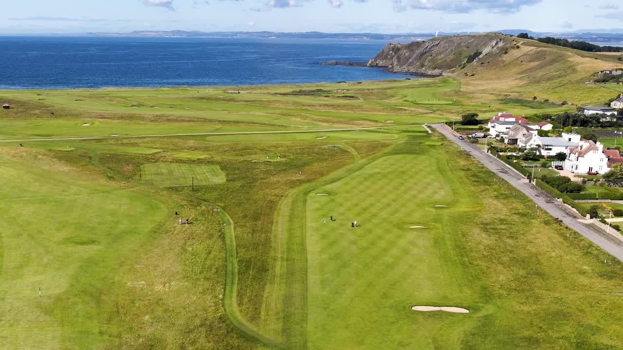 Drone glides above scenic golf course, coastline, and village under bright daylight, steady aerial movement