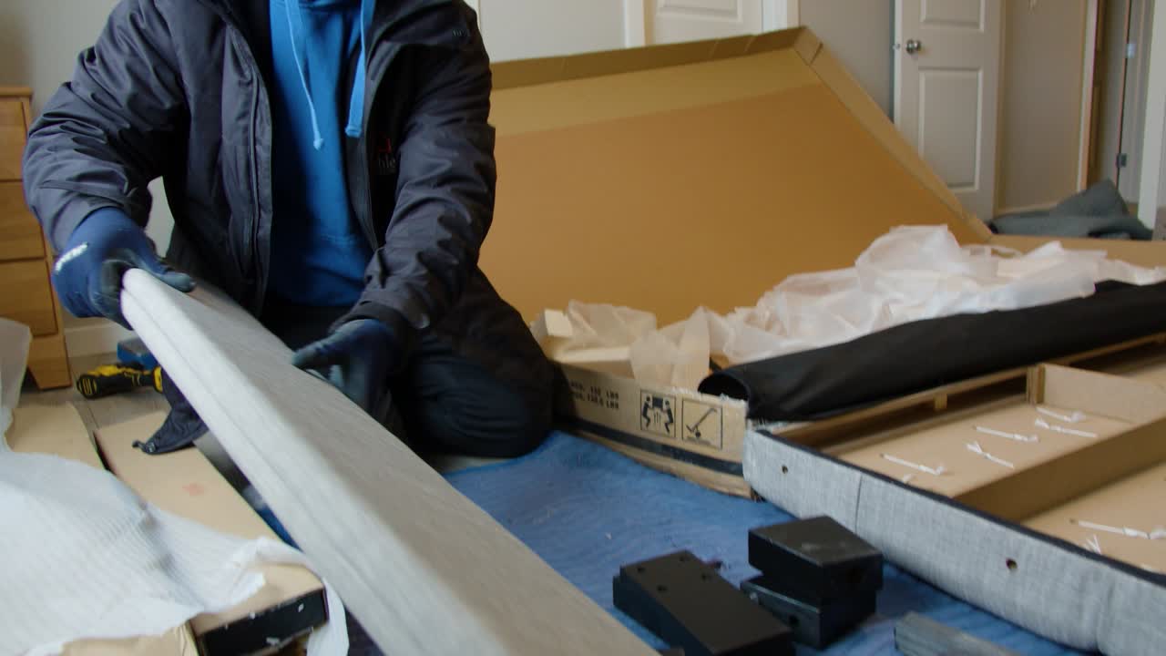 Action shot of delivery worker with gloves unpacking bed frame parts from large box inside customers home