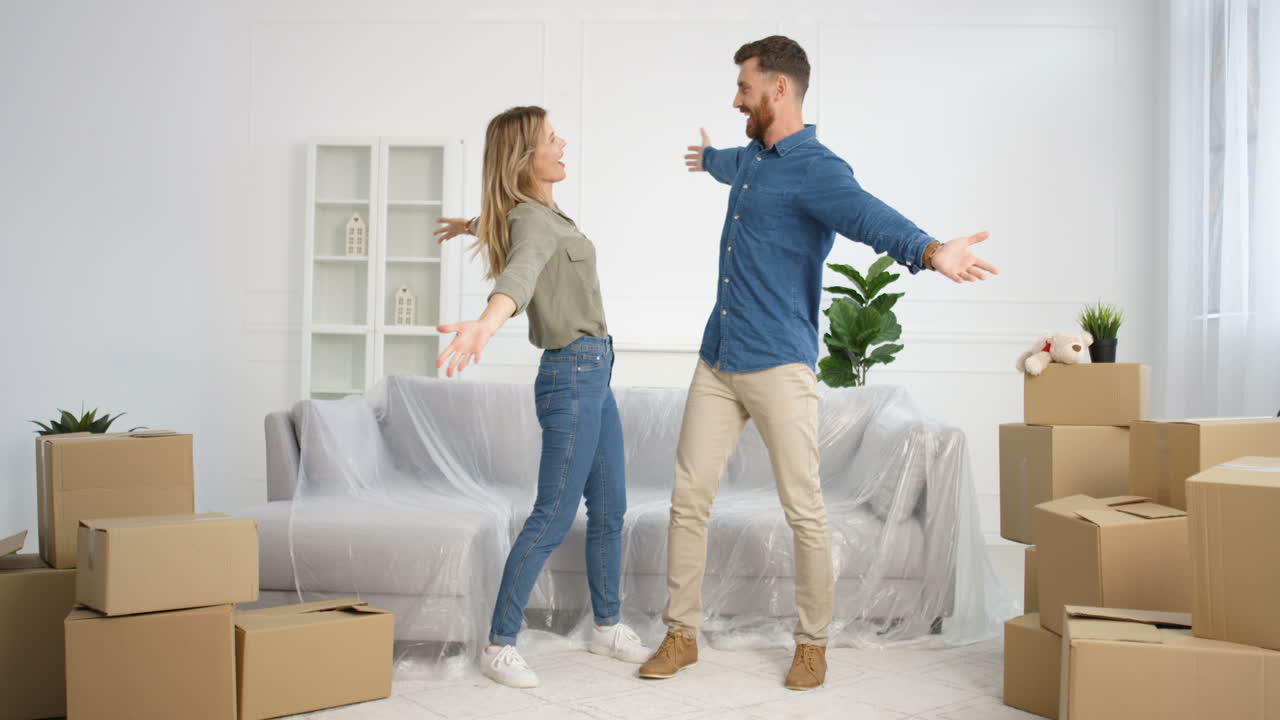Caucasian young cheerful married couple having fun, laughing and jumping in living room among carton boxes