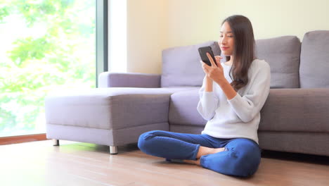 젊은 이국적인 여성이 집 바닥에 앉아 웃고 생각하면서 스마트폰으로 콘텐츠를 스와이프하고 풀 프레임 슬로우 모션