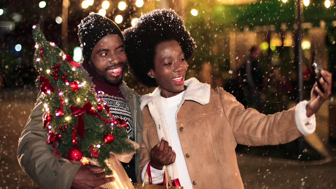 vista de cerca de una alegre pareja afroamericana haciéndose un selfie con un smartphone mientras nieva en la calle en navidad
