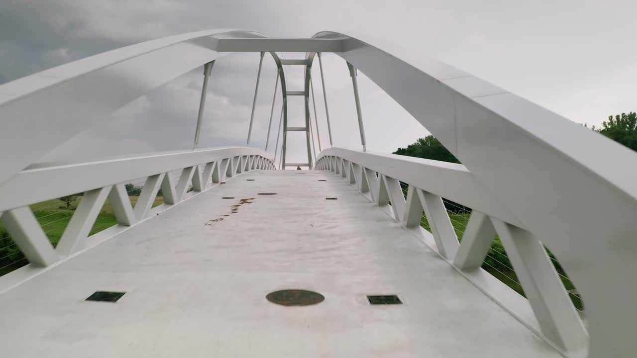 Flying forward through a cycling bridge running across calm river Moravia in Slovakia revealing a person walking towards the camera. Aerial sunset footage taken just before a storm