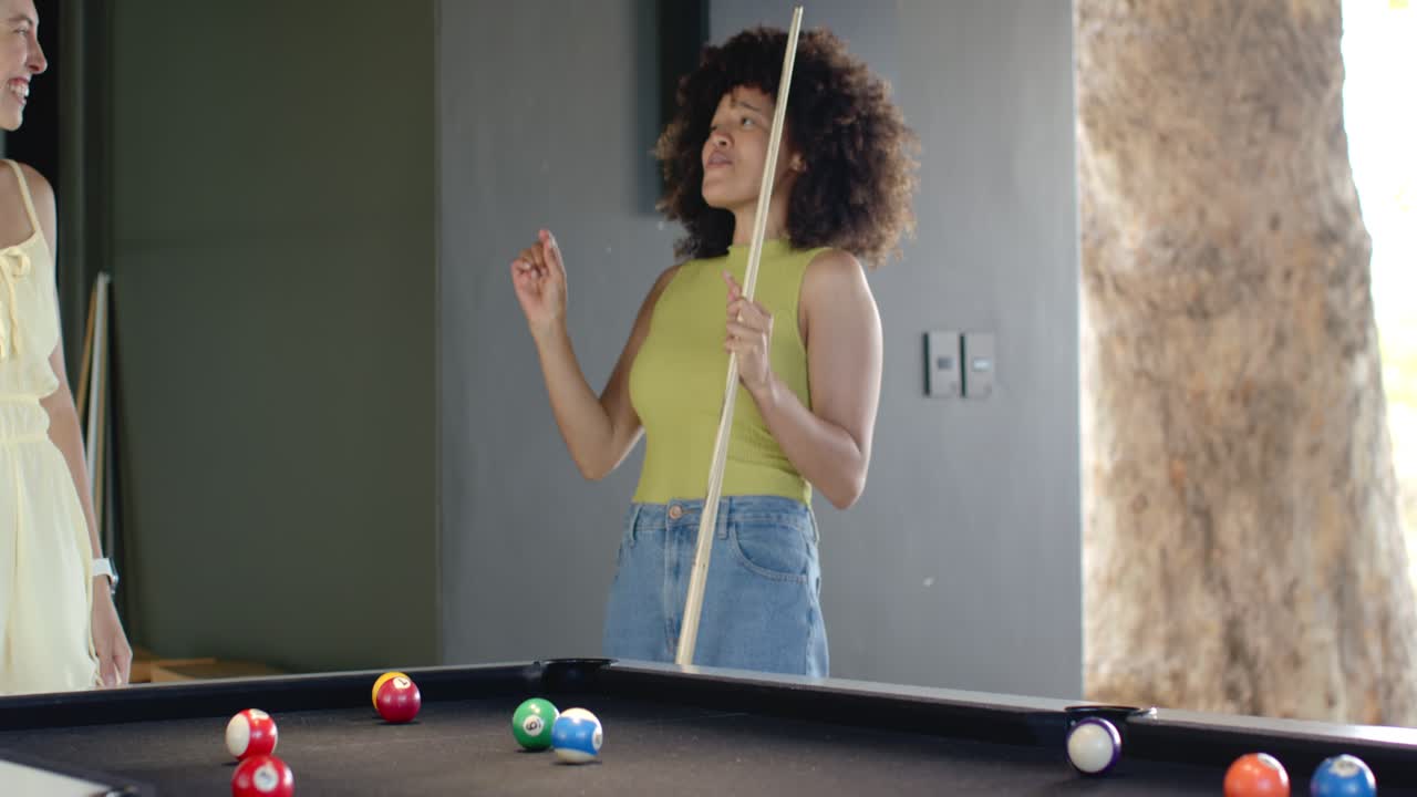 Diverse female friends, African American woman in lime top aiming, striking cue at home pool table