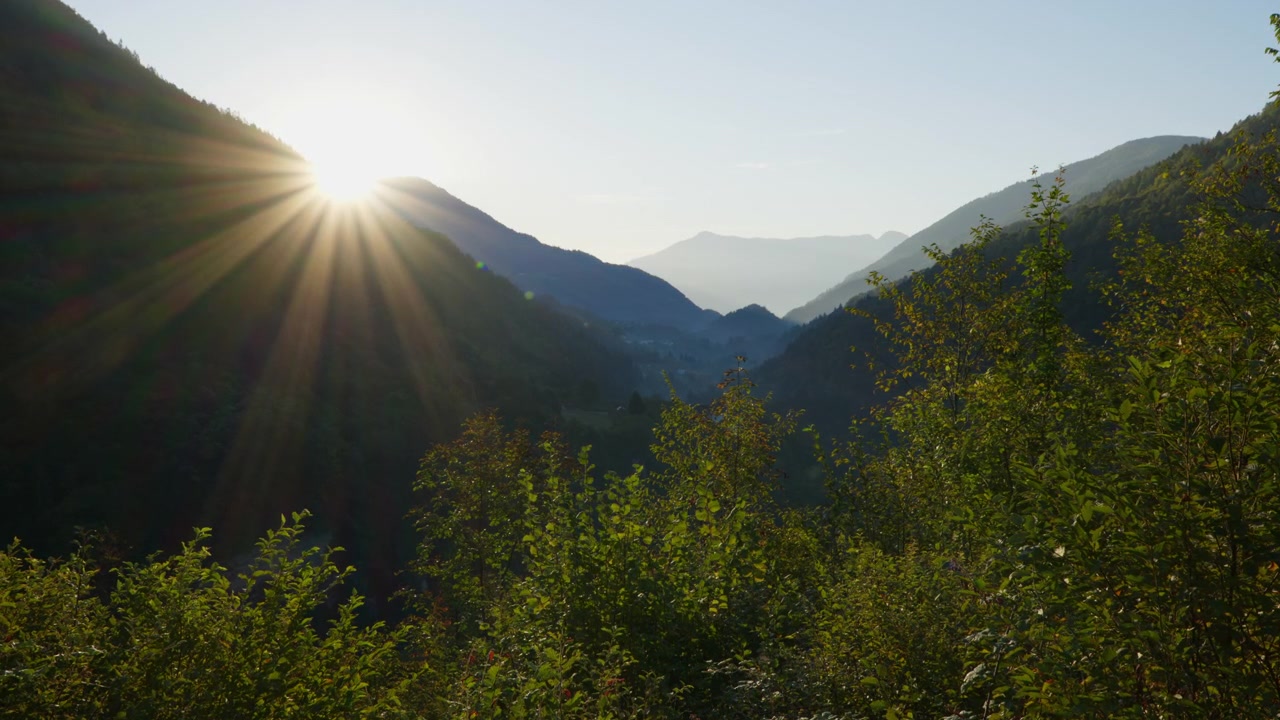 Low sun bursts over a narrow Italian valley, sending rays across forested hills and green shrubs in the foreground.