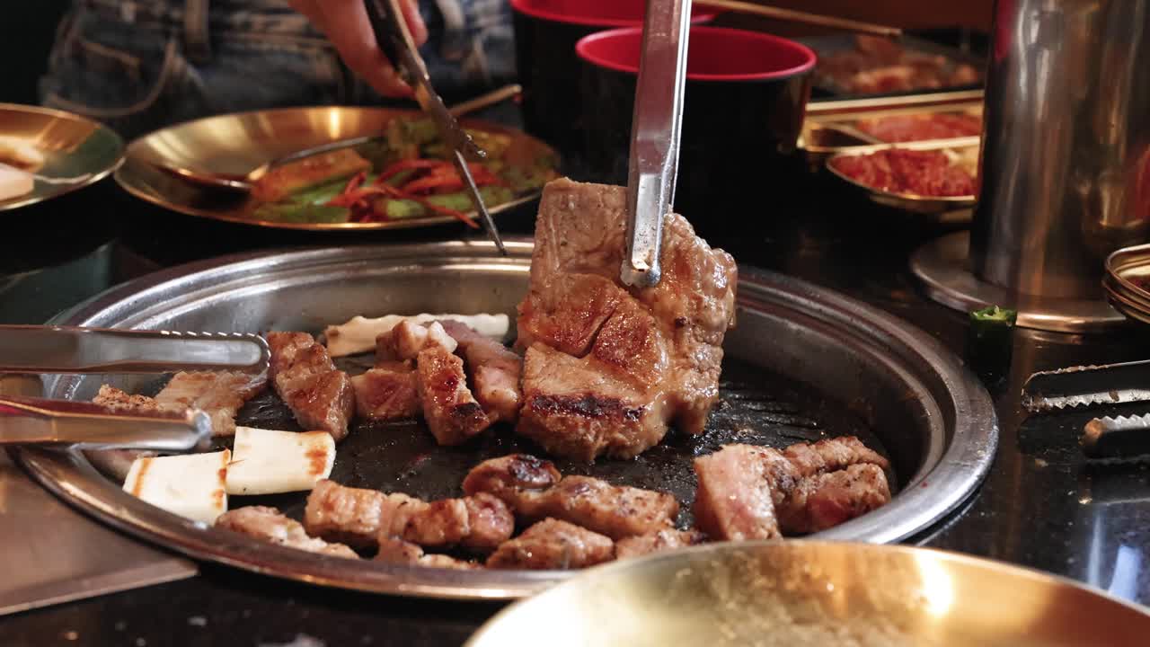 Hand uses tongs and scissors to cut pork belly on tabletop grill, warm ambient lighting