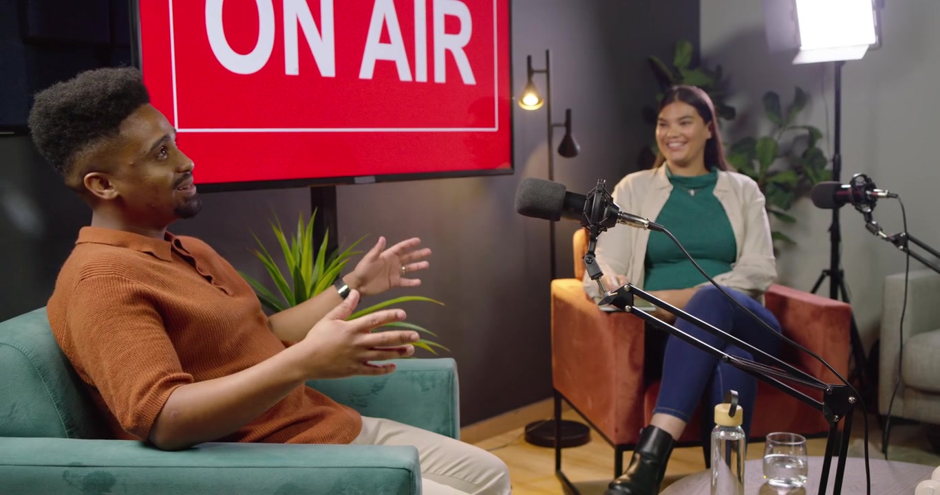 Uomo e donna afroamericani che registrano un podcast, un ospite alza la mano e parla, l'uomo gesticola vicino alla scritta ON AIR.