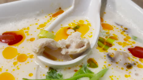 authentic and delicious tom kha gai thai with chili oil and herbs in white bowl and large spoon