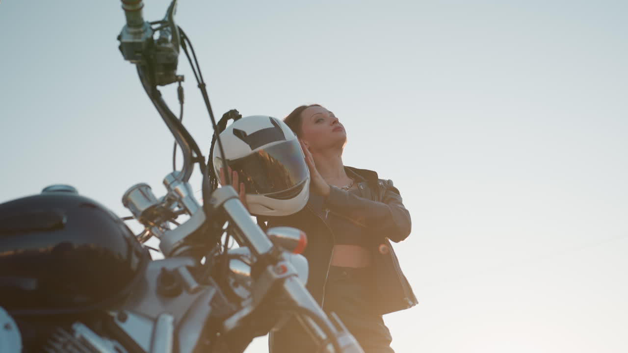 Elegant lady removes helmet with confident pose wearing leather jacket standing near motorcycle under daylight showing bold attitude determination and strength while city background
