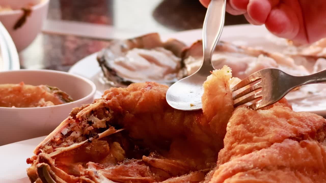 Close-up of hands using fork and spoon to enjoy crispy fried fish with side dishes.
