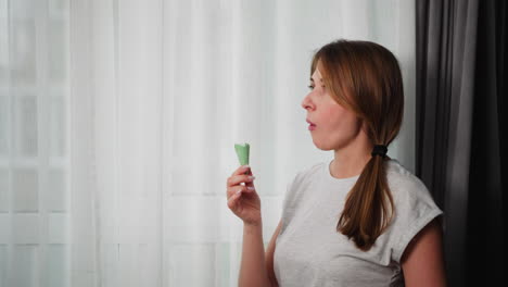 side view of woman with brown hair tied in ponytail wearing grey top enjoying ice cream cone with green waffle near sheer curtain in bright cozy interior, savoring calm and refreshing moment indoors