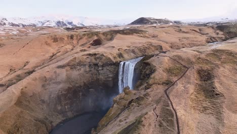 drone view of iceland’s skógafoss waterfall from afar, surrounded by golden hills and snowy mountains.