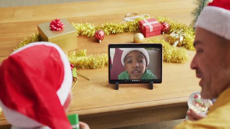 padre y hijo caucásicos con sombreros de santa usando una tableta para una videollamada de navidad con el niño en la pantalla