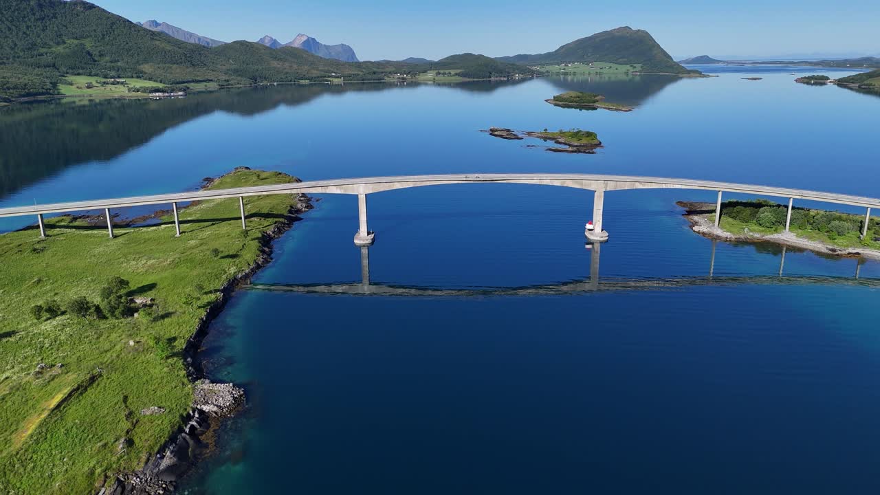 Drone footage flying over a bridge on Engeløya island in northern Norway, with two cyclists enjoying the early morning view of calm water and mountains