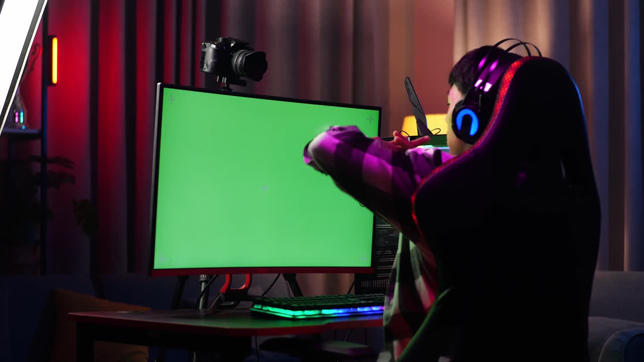 Back View Of Asian Boy Streamer Stretching After Using Green Screen Personal Computer. Live Stream On Desk Illuminated By Rgb Led Strip Light