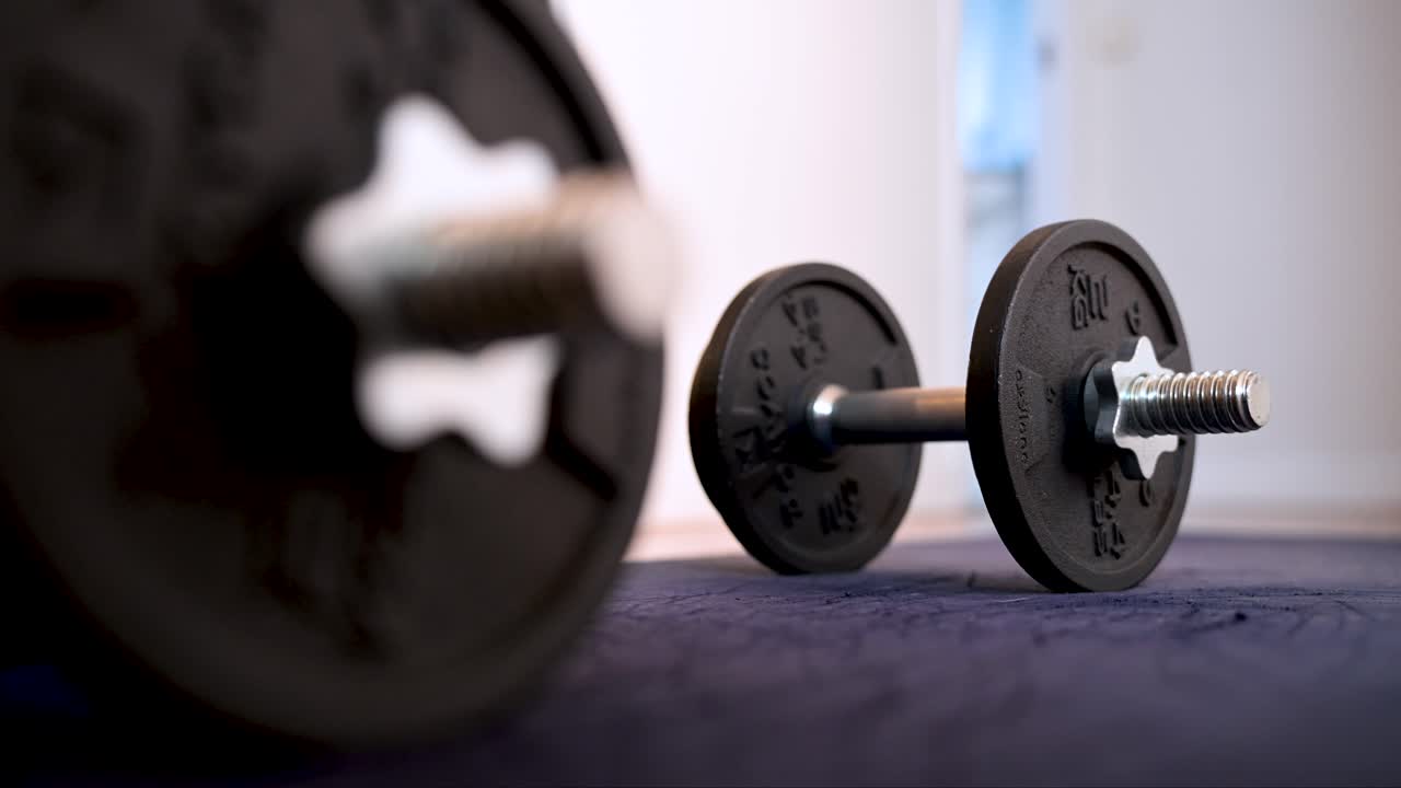 A rack focus shot between two dumbbells lying on a yoga mat. A cinematic close-up of free weights for a home workout or strength training session