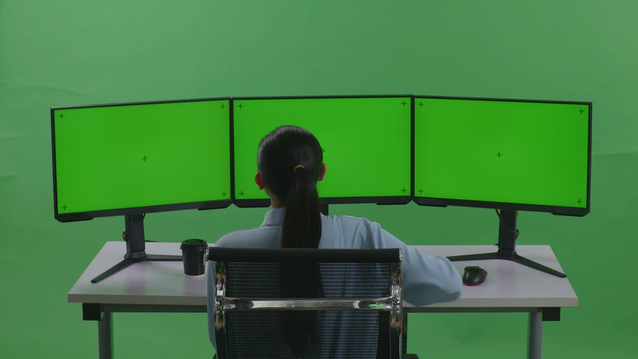 vista trasera de una mujer asiática con dolor de espalda mientras trabaja con múltiples monitores de computadora simulados en un estudio de pantalla verde