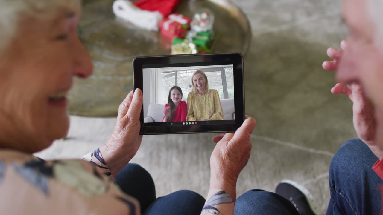 pareja caucásica mayor sonriendo y usando una tableta para una videollamada de navidad con la familia en la pantalla