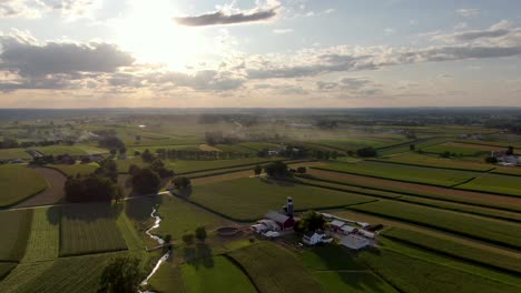 Gorgeous-descending-aerial-of-Amish-farmland-in-Lancaster-County,-Pennsylvania,-during-summer-sunset,-creek-bisects-colorful-fields-making-patterns