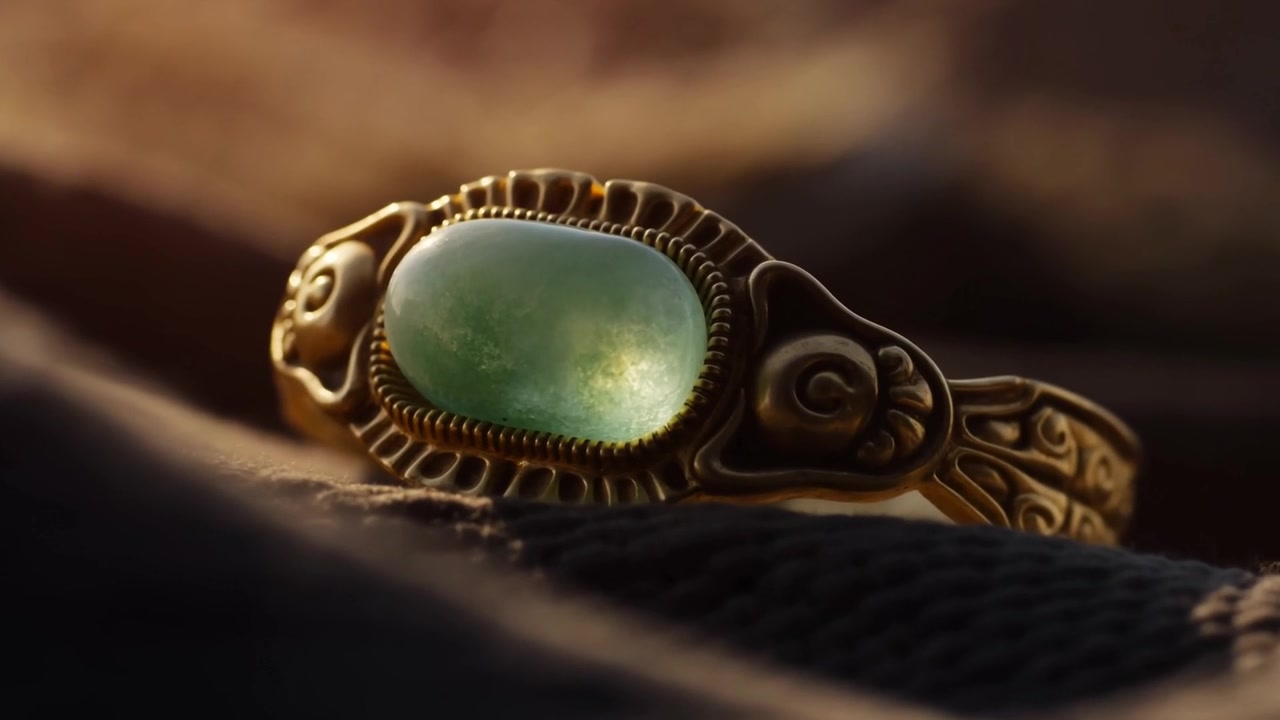 Ajustando o foco, anel de ouro revela cabochão verde e aro cravejado sobre tecido dobrado com luz quente.