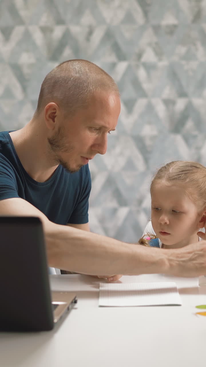 concept of distance learning of father and little student daughter at white table in room of house, with laptop, scissors, book, dad shows how to correctly paste colored red circle