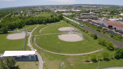 Vista-Aérea-Del-Tren-De-Cercanías-Pasa-Por-Un-Campo-De-Béisbol-Vacío-En-Un-Día-Soleado-De-Verano