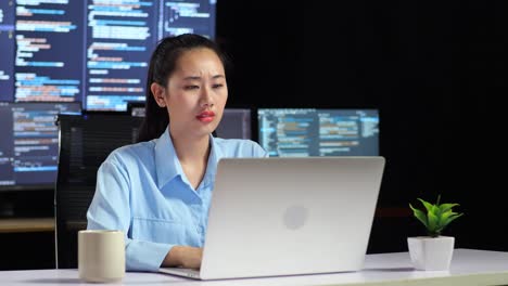 asian female programmer thinking about something while writing code by a laptop using multiple monitors showing database on terminal window desktops in the office