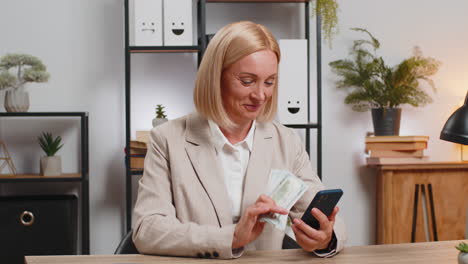 mature businesswoman counting money dollar cash using smartphone calculator app at home office desk