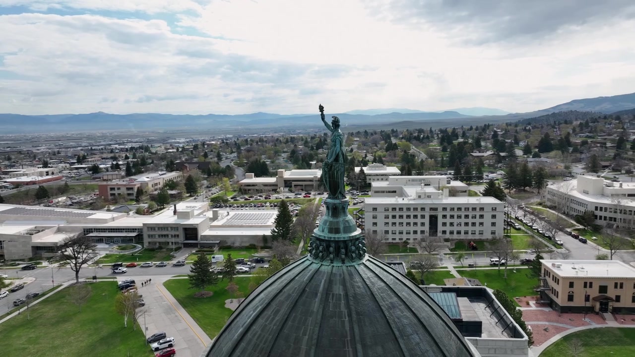 Statua in verderame che regge una torcia in cima alla cupola del Campidoglio, che domina i tetti di Helena, i parcheggi e un moderno complesso di uffici sotto una catena montuosa nebbiosa. Helena, Montana, USA. Ripresa aerea orbitale con drone