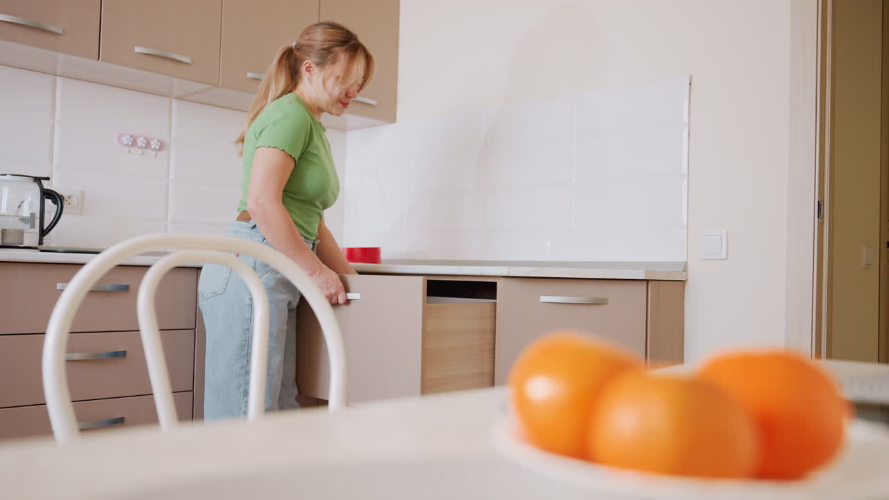 Donna con camicia verde e jeans ripone una padella in un cassetto mentre si trova in una cucina luminosa; il piano di lavoro è visibile, con una ciotola di arance in primo piano, creando un'atmosfera di vita domestica informale