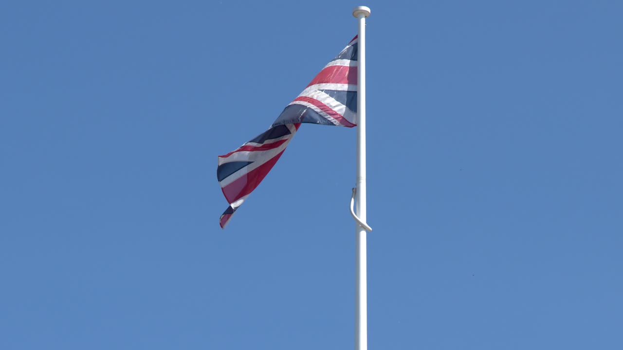 Union Jack flag waves atop historic architecture under clear blue sky, midday sunlight, static camera